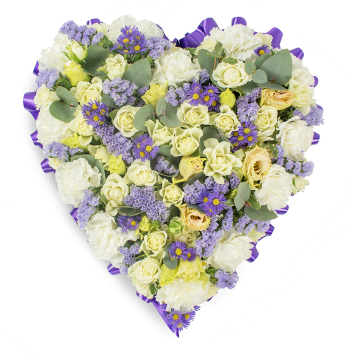 A tender, heart-shaped sympathy wreath presented on a pristine white studio surface, its shape crisply defined by a continuous band of saturated purple satin ribbon that creates a graceful frame for the flowers within. The interior is a dense, harmonious blend of soft whites, creams and pale yellows: compact cream and pale yellow roses form the primary texture, their layered petals offering a plush, intimate surface that moves from closed buds to gently opening blossoms. Complementing the roses are clusters of classic white carnations, their ruffled faces adding softness and traditional comfort. Threaded through this pale foundation are feathery sprays of lavender-hued statice, lending an ephemeral, meadow-like quality, and small purple daisy-like asters with bright yellow centers, which give focal contrast and a subtle lift to the composition. Blue-green eucalyptus leaves, rounded and slightly silvery, are inserted at intervals to break the floral mass and introduce a cool, aromatic note that suggests a faint, calming scent. The overall palette and tactile layering create a serene, contemplative aesthetic ideal for memorial services, wakes, or graveside placement. Lit evenly to honour the arrangement's quiet dignity, this piece reflects the skill of a local florist who understands how to express condolence through colour, form and texture - a design that would sit respectfully in chapels or near the peaceful corners of Woodside's community gathering places.