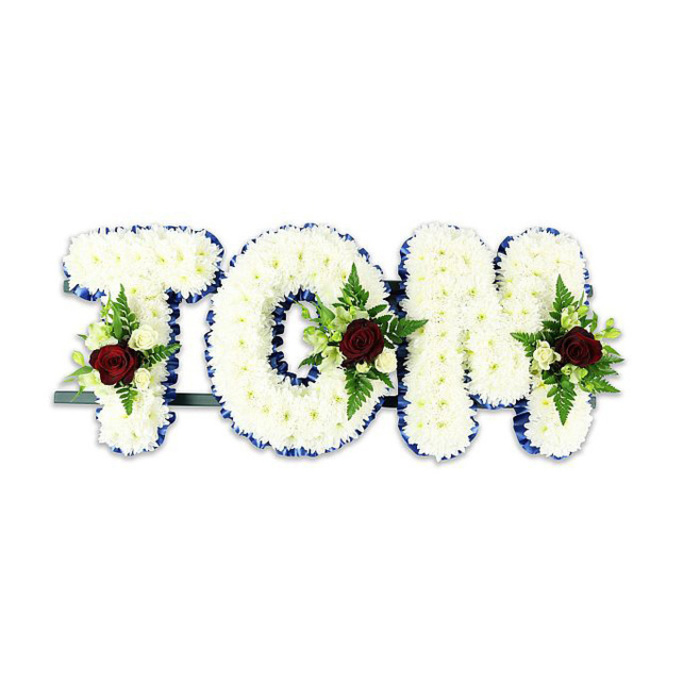 An intimate, respectful floral tribute spells out TOM in large, sculpted letters, each letter meticulously covered in a dense layer of small white chrysanthemum blooms that read as soft, pillowy texture and catch light with a satin-like glow. A striking royal blue ribbon traces the perimeter of every letter, creating a formal outline that separates the white surface from its surroundings and gives the composition a composed, ceremonial air. At the heart of each letter a single, rich red rose commands attention; its velvety depths are framed by petite creamy-white spray roses and delicate fern fronds that introduce fresh green accents and gentle movement. The letters are subtly anchored by a slim dark green or black metal support visible along the base, designed to keep the tribute steady when displayed upright in a chapel, funeral home or graveside service. Set against a stark white background in the image, the contrast between white chrysanthemums, royal blue ribbon and crimson focal roses becomes quietly powerful-measured, dignified and consoling. The arrangement carries a restrained fragrance, a clean, green note when one draws near, and the overall presentation suggests the hands of an experienced florist who understands how to balance texture, colour and symbolism. This format of named floral tribute is commonly chosen by families in the Woodside area who wish to mark a life with something personalised and timeless; Florist Woodside often crafts similar bespoke memorial letters for local services, tailoring the spray details to reflect individual sentiment while ensuring the piece reads clearly from a distance.