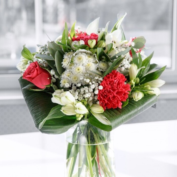 A classic yet modern bouquet in deep red and crisp white stands in a clear cylindrical vase, the stems neatly cut and fanned out in the water like a carefully planned design sketch. It feels very much like the kind of arrangement a Woodside family might place on the hall table to welcome guests, full of colour and quiet ceremony. At the core, several deep red roses glow with a velvety richness, their petals curling outward in perfect spirals that suggest enduring love. A large crimson carnation, heavily ruffled and full-bodied, presses forward at the front of the bouquet, lending a sense of movement and texture, almost like a piece of finely gathered fabric. Surrounding these bold red flowers are soft white blooms: alstroemeria with long, slender petals marked with subtle speckles, rounded daisy-like flowers, and several white lily buds still closed, their sleek forms hinting at elegant flowers yet to reveal themselves. Fine clouds of baby's breath, or gypsophila, float throughout the arrangement, their tiny white stars filling gaps and casting a filmy, romantic veil over the whole piece. Lush greenery frames everything: broad, glossy dark leaves are artfully wound around the base to create a structured collar, while longer, slightly bluish eucalyptus-style stems and slim pointed leaves rise up to add height and contrast. The bouquet is placed on a pristine white surface, with a bright, softly blurred background that suggests daylight filtering through a nearby window, perhaps on a quiet street off Woodside's main road. The mood is uplifting and affectionate, equally suited to a thoughtful anniversary surprise, a Mother's Day delivery to a home near Woodside Green, or a graceful thank-you gift that brings a polished burst of nature into an everyday room.