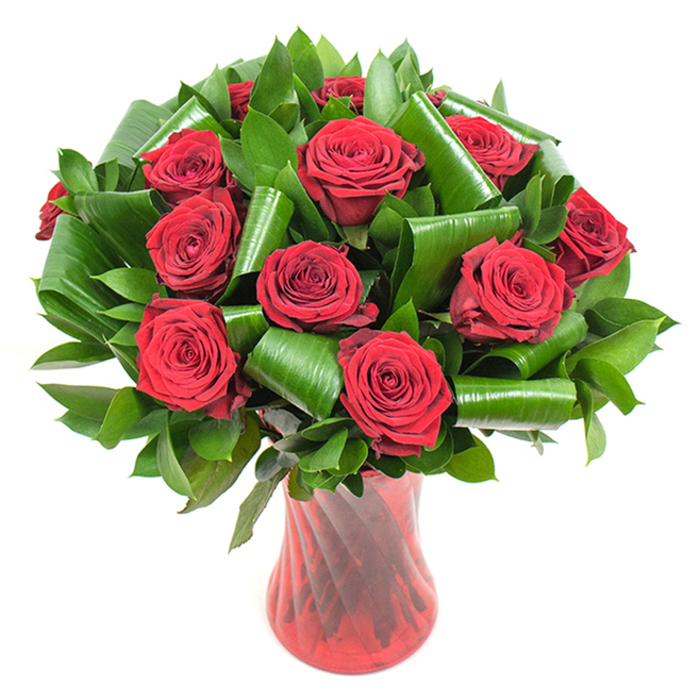 This photograph presents a densely packed, rounded bouquet of deep crimson roses displayed with intentional elegance: each rose is in peak bloom, its velvety petals layered into concentric spirals that suggest both freshness and romance. The intense red is offset by a generous quantity of dark green foliage - smaller glossy leaves build a verdant, textured backdrop while larger emerald leaves have been gently rolled and folded into sculptural coils that frame and cradle the roses. Those curled leaves create striking arcs and negative space, lending a contemporary, architectural sensibility to a very traditional flower. The arrangement is held in a slender, ruby-red glass vase whose translucent surface features subtle twisted ribs; these vertical lines catch the light, sending reflective highlights that harmonise with the roses' tones and give the piece a refined vertical energy. Photographed against a pure white background, the bouquet's colours and textures are isolated and amplified so that the eye rests on the tactile contrast between soft petals and structured greenery. The overall mood is one of considered passion and quiet luxury - a bouquet tailored for an important anniversary, a Valentine's gesture, or a thoughtful celebration that calls for classic romance with a modern twist. As a florist familiar with Woodside and its leafy pockets near Woodside Green and the local High Street, I imagine this arrangement arriving carefully boxed for a neighbour awaiting flowers, its fragrance spilling out on unwrapping: a warm, familiar scent that suggests comfort, devotion, and attention to detail.