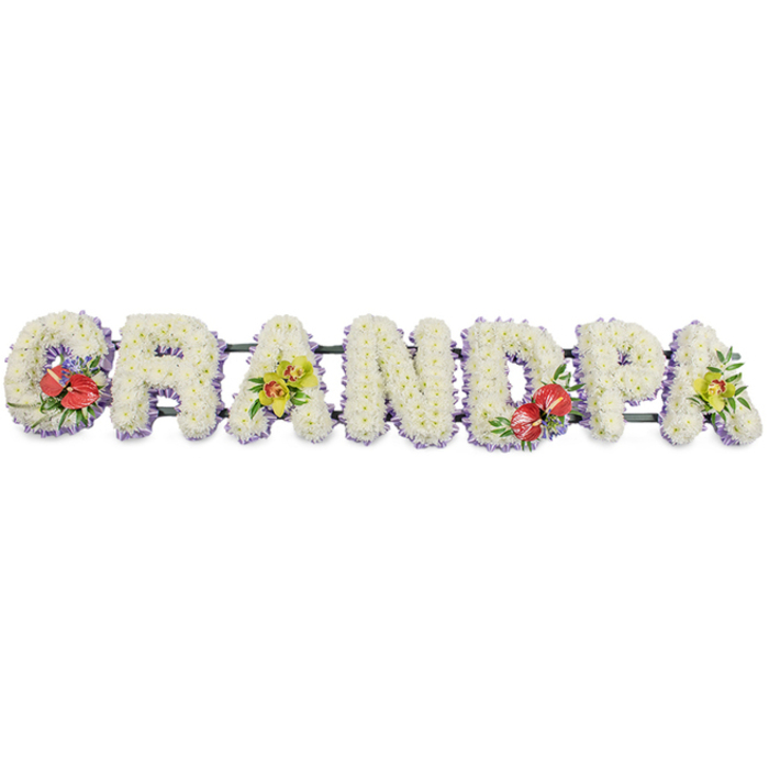 The image shows a carefully crafted funeral flower tribute spelling "GRANDPA" in large, upright, block capitals, stretching horizontally across a plain white field so the eye rests wholly on the flowers themselves. Each letter is sculpted from dozens of small, pristine white chrysanthemums or similar cushion blooms, their rounded heads tightly massed together to create a thick, almost quilted surface that radiates purity and a sense of gentle rest. Along the lower edges of the letters runs a slim trim of pale lilac or lavender ribbon, its cool hue subtly outlining the shapes and adding a whisper of soft colour reminiscent of evening light over Woodside's quiet streets. Supporting bars in dark green run along the base, mostly hidden by the floral work but providing the strong, practical framework needed for display on an easel, hearse, or at a graveside. Carefully positioned sprays of accent flowers break the expanse of white and draw attention to particular letters. On the "G" and the "D", rich red anthuriums stand out dramatically, their glossy, heart-shaped spathes and clear yellow spadices framed by polished green leaves and a scattering of fine filler blooms, suggesting heartfelt passion and enduring love. On the first "A" and the last "A", clusters of yellow-green orchids cascade gracefully, their delicate, sculpted petals and unusual colouring adding a sophisticated, almost timeless note, nestled among lush foliage. The tribute feels composed for a beloved grandfather, perhaps travelling from a florist near Woodside Green to a funeral service or memorial in the local area. Every element - the clean typography, the white flowers, the refined accents - works together to convey respect, gratitude, and a tender farewell that feels both personal and dignified.