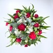 A lush, circular bouquet is photographed from directly above against a clean white background, every stem gathered so tightly that the flowers seem to spill toward the viewer. At the centre, velvety deep red roses unfold in varying stages of bloom, from tight buds with curled outer petals to more open blossoms that reveal densely layered hearts, conveying romance and quiet intensity. Around them, ruffled fuchsia and ruby red carnations create a soft, almost cloud-like texture, their frilled edges catching the light and adding a joyful, celebratory feel. Clusters of dainty white spray chrysanthemums and other small, daisy-like white flowers glimmer between the stronger reds, offering cool contrast and giving the composition a fresh, breathable brightness. Fine sprigs of white gypsophila, or baby's breath, appear like scattered stars, threading delicately through the bouquet and softening any hard lines. Broad, glossy dark green leaves, likely from lily or alstroemeria stems, radiate outward to give the arrangement its structure, while rounded, silvery-green eucalyptus leaves weave in and out, their muted tone and slightly waxy surface bringing a contemporary edge to a classic romantic palette. The lighting is even and natural, with no shadows to distract from the details, making every petal and leaf appear crisp and newly arranged. The bouquet feels full of movement and life, as if just tied on a workbench in a Woodside florist's studio before being sent out for same-day delivery across the area. It carries the mood of a heartfelt embrace-ideal for anniversaries, Valentine's surprises, birthday celebrations along local streets, or a vibrant "thinking of you" message that brings warmth into a Woodside home.
