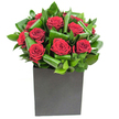There is a magnetic simplicity to this arrangement: a compact cluster of deep crimson roses whose velvety petals unfurl in soft, generous spirals at the heart of the design. Each bloom sits slightly turned to catch the light, their rich red tones intensified by the glossy, sculptural leaves that have been gently curled and layered around them. The foliage creates a rhythmic, overlapping pattern of vibrant green that adds both movement and depth, like waves framing a central flame. The composition rests in a crisp, matte black cube - a box vase whose squared lines and grounded finish contrast the lushness above and lend the whole piece a modern, urban poise. Lighting in the photograph is clear and steady, picking out the sheen on each leaf and the velvet gleam of the rose petals so the arrangement reads as both neat and emotive.

Brought to life with careful floral technique, the tactile contrast between silken petals and the firmer, almost waxy texture of the greenery invites you to imagine the scent: a clean, classic rose perfume brushed with fresh green notes, bright and slightly tropical. As a local florist might advise, this bouquet is composed with intention - elegant enough for an anniversary, bold enough for a milestone celebration, and intimate enough to convey deep gratitude. It also fits contemporary interiors, from a Woodside living room to a compact office near the tram stop.

Designed to make a personal statement, the piece reads as a quiet declaration of affection and style, a memorable gift that feels both polished and heartfelt for recipients across Woodside and neighbouring South Norwood.